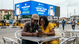 A young  man and woman sitting together at a small table outside, both looking at a smartphone screen with expressions of excitement
