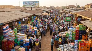 An open-air market scene, with traders selling packaged consumer goods, beverages, and household products on display at their stalls.