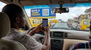 A man in a car stuck in traffic, holding a phone and scanning a QR code on a large billboard